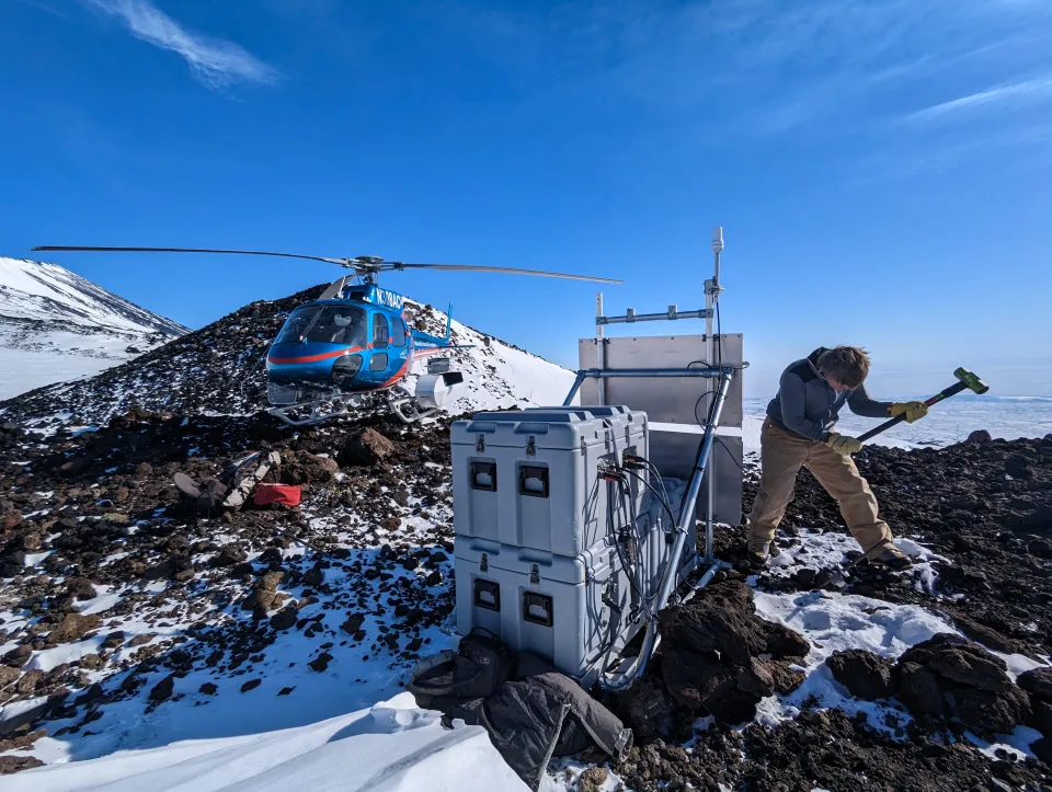 Helicopter next to instruments installed in Antarctica, with an engineer swinging a hammer.